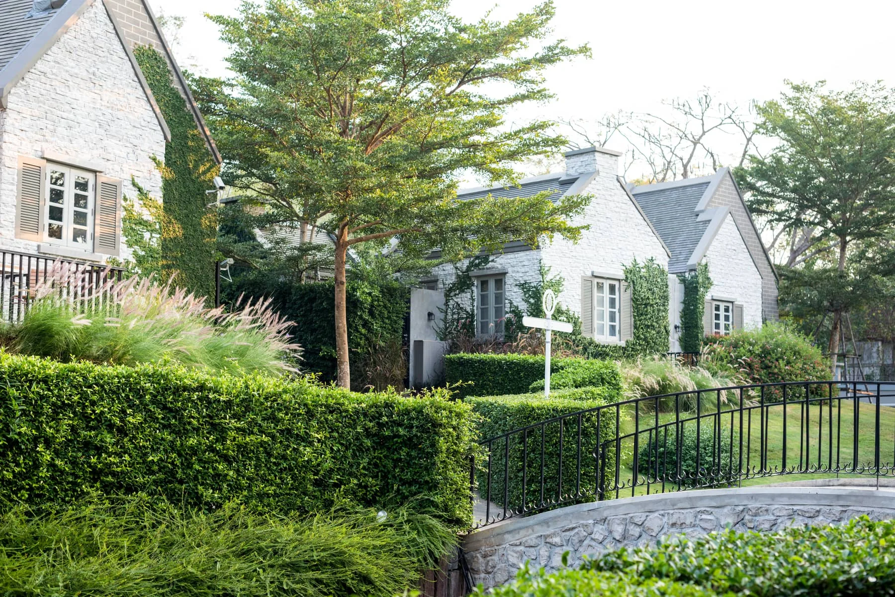 Stone and stucco house behind a trimmed hedge, an iron fence and a landscaped garden