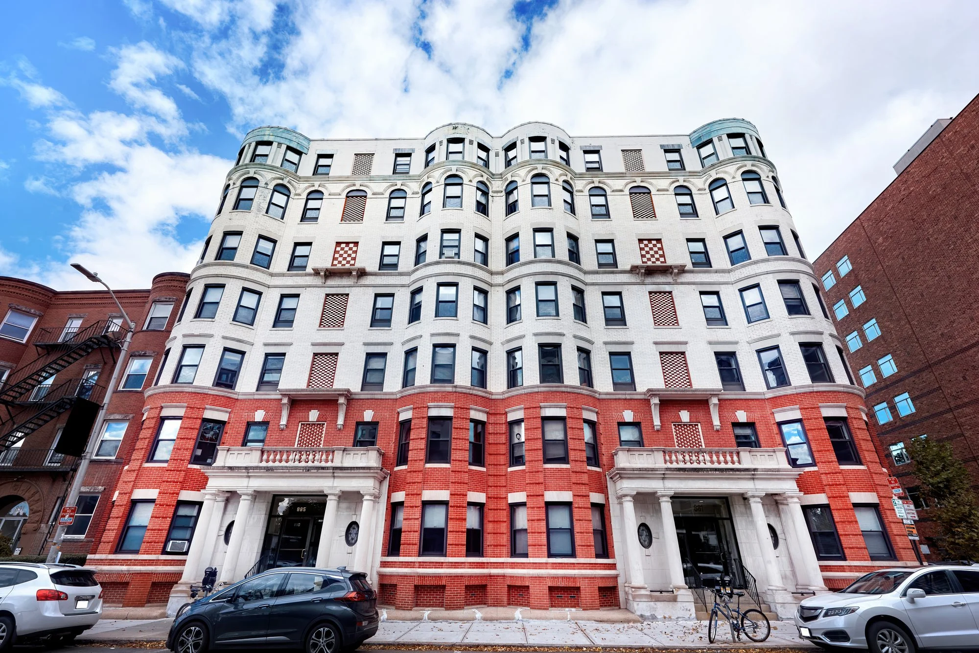Tall red-brick and white apartment building with arched entrances seen from street level under a blue sky