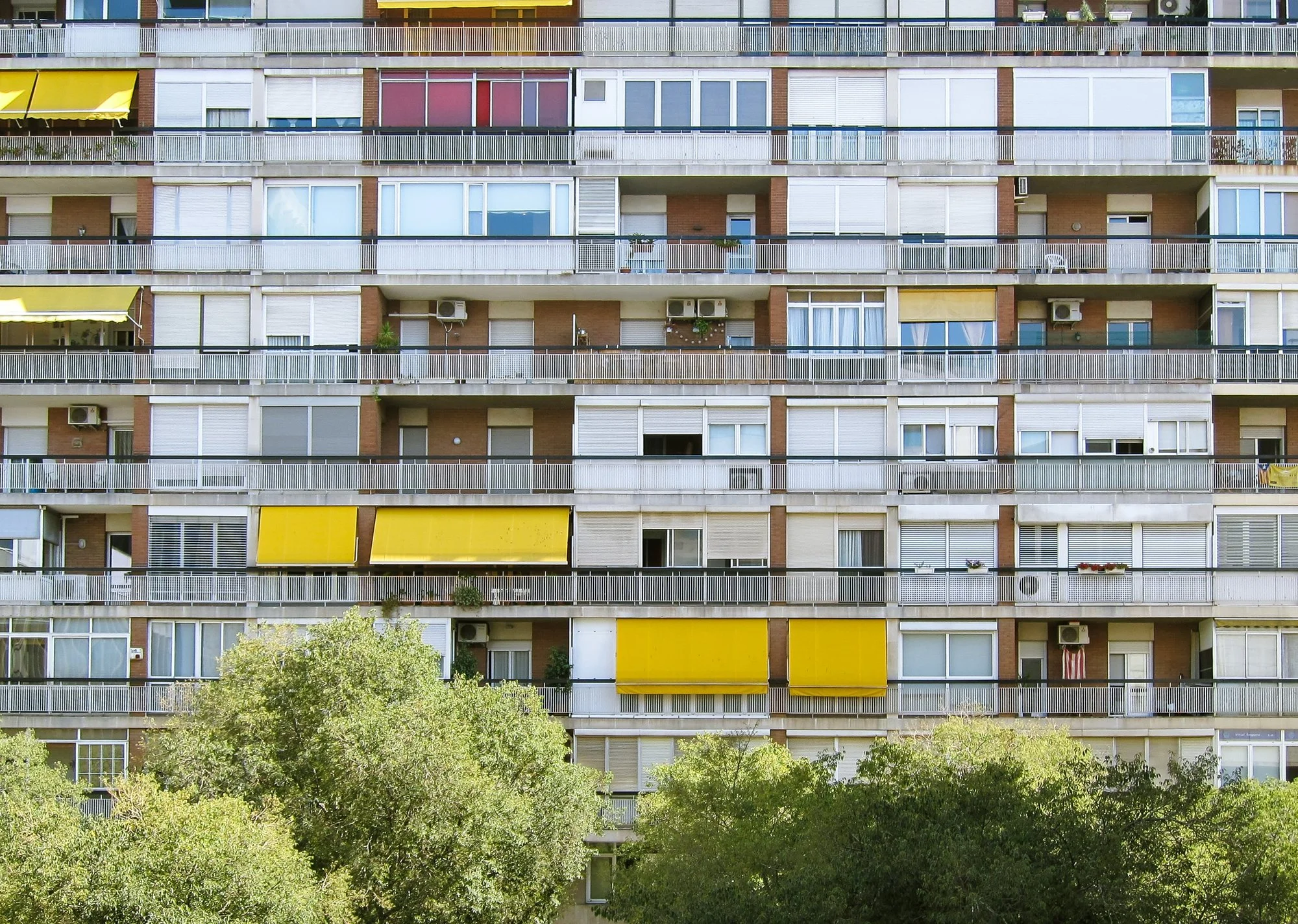 Long mid-rise apartment building with rows of balconies and yellow awnings behind green trees