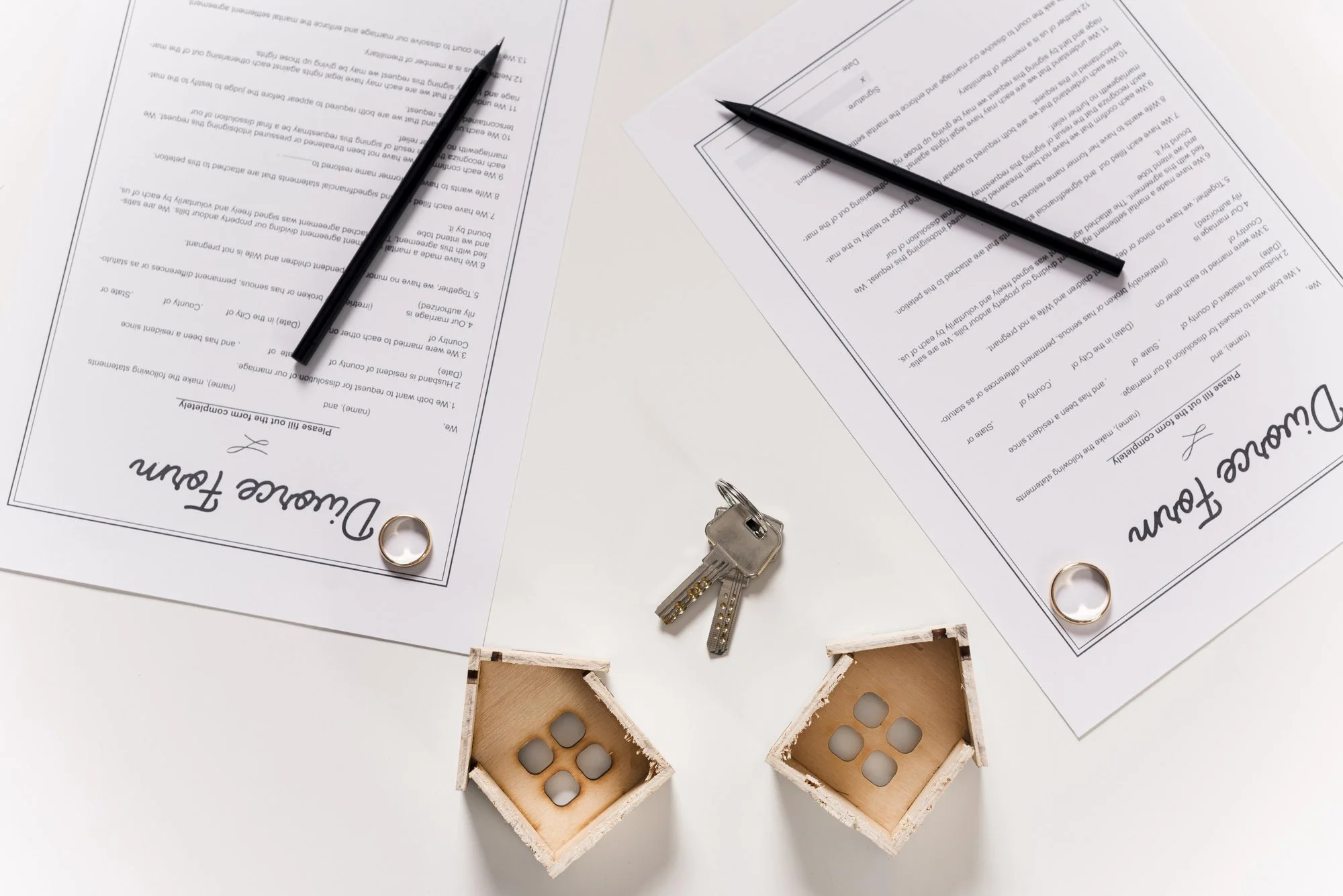 Two divorce forms with pens, wedding rings, house keys and small wooden houses on a white table