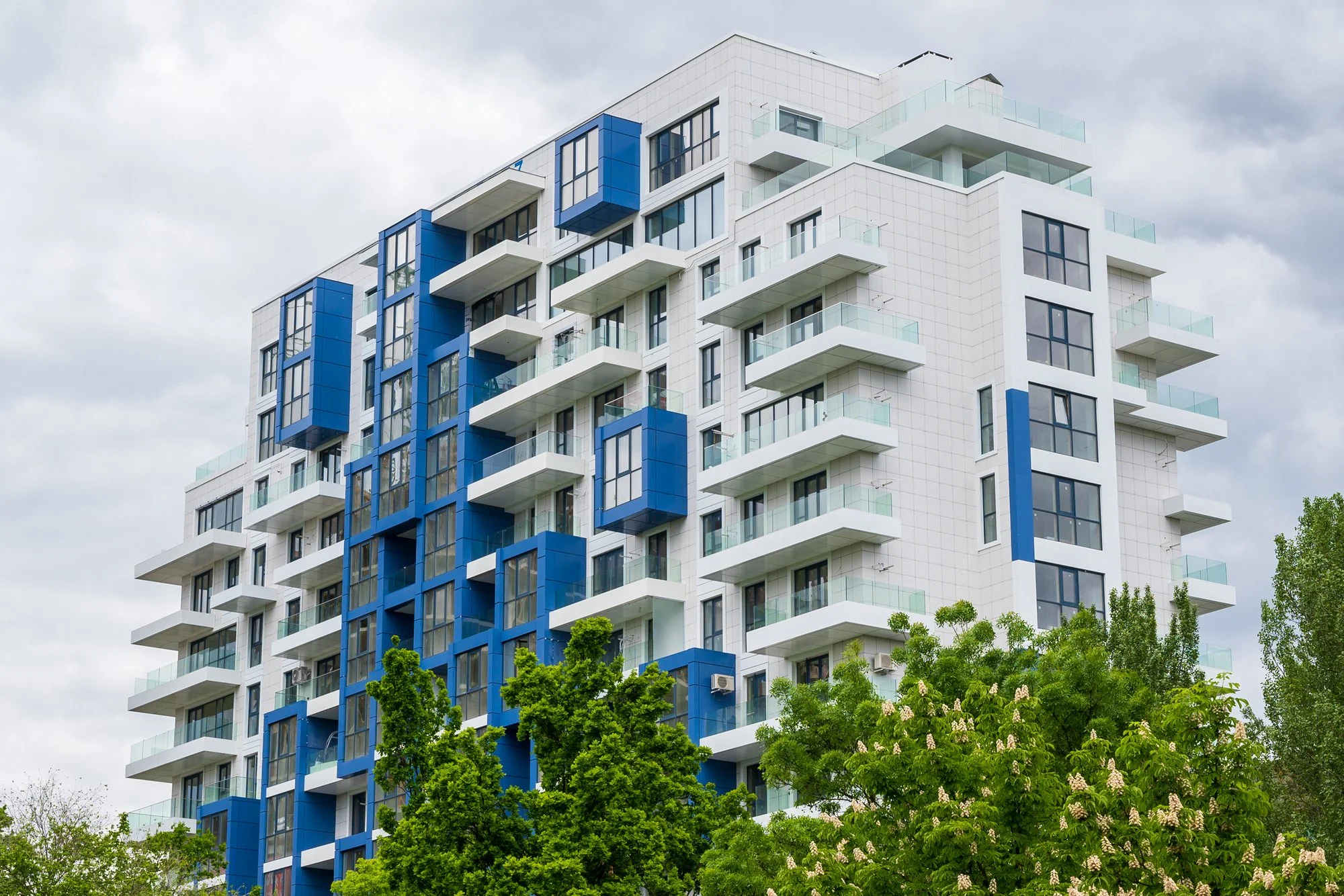 Modern white apartment building with blue balcony details behind green trees
