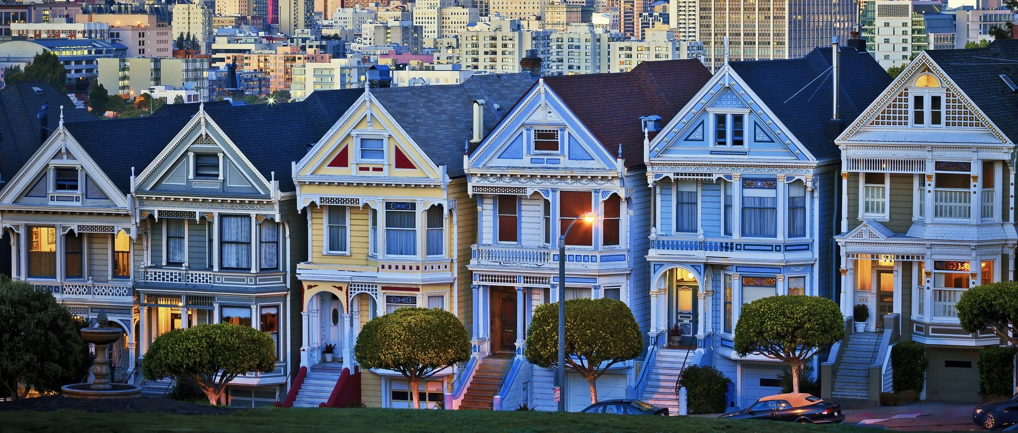 Row of colorful Victorian houses with a city skyline behind them at dusk